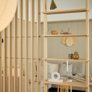 WoodUpp Raumteiler-Regal aus hellem Eichenholz: schlanke vertikale Lamellen verbunden mit schlanken Metallstangen und seitlichen, offenen Regalböden. Das freistehende, minimalistische Möbel zeigt mehrere Dekostücke (Holzschale, Glasskegel, Metallbecher, Marmorring, Kerze, birnenförmige Holzfigur), dezentes Licht, vor einem gemütlichen Sofa und schafft warmes, skandinavisches Wohnambiente.