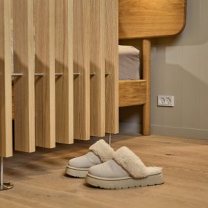 WoodUpp zeigt ein warmes Schlafzimmer-Detail: im Vordergrund ein helles Holz-Lamellenpaneel von WoodUpp mit sichtbaren Metallstegen und stabilen Füßen, dahinter ein schlichtes WoodUpp-Bettgestell mit Kopfteil und Matratze in passendem Holzton. Auf dem Holzboden liegen flauschige, cremefarbene Hausschuhe neben einer weißen Steckdose.