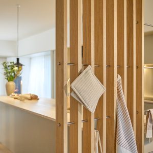 WoodUpp Lamellenwand aus hellem Eichenholz als dekorativer Raumteiler; in den senkrechten Latten sind schlanke, polierte Edelstahlhaken und -stangen von WoodUpp eingesetzt, die als praktische Aufhängung für Topflappen, Geschirrtücher und Küchenutensilien dienen. Im Hintergrund Kücheninsel mit Vase, Kerze und warmem Licht.