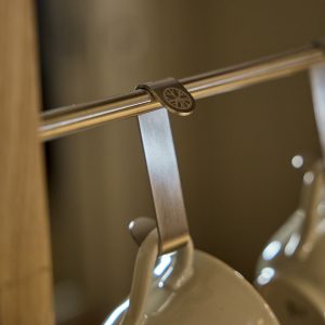Detailaufnahme eines WoodUpp Edelstahl-Tassenhakens: ein gebürsteter Metall-Clip mit eingraviertem WoodUpp-Logo, aufgelegt auf einer chromglänzenden Stange und trägt eine helle Keramiktasse. Produkt wirkt schlank, mattiert und stabil, passend für Küchenstangen zur ordentlichen Aufbewahrung von Bechern.