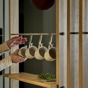 WoodUpp Holzregal in heller Eiche mit offenem Fach und schmaler Ablage, ausgestattet mit einer eingebauten, matt gebürsteten Edelstahl-Hakenleiste von WoodUpp, an der fünf gleichmäßig hängende weiße Keramiktassen befestigt sind; zwei Hände nehmen eine Tasse, auf der Ablage liegt ein frischer Rosmarinzweig.