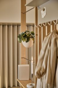 Foto einer WoodUpp-Wandgarderobe aus hellem Eichenholz mit schmalen Vertikalleisten, Regalböden und Metallstangen. Zu sehen sind ein hängender halbrunder weißer Pflanzkübel, Bücher auf dem Regal, ein schlanker Kerzenhalter mit marmoriertem Fuß und ein Mantel an Haken — minimalistisches WoodUpp-Design sowie warmes Kerzenlicht und dezenten Vorhängen im Hintergrund.