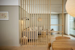 Wohnraum mit WoodUpp Holzlamellen-Raumteiler und integriertem WoodUpp Regal: schlanke helle Eichenholz-Lamellen mit quer verlaufenden Metallstäben, freistehend zwischen Ess- und Wohnbereich. Im Hintergrund graues Sofa, Rattan-Beistelltisch, Teppich, Körbe, runder Holzesstisch im Vordergrund, zarte Vorhänge und warmes Tageslicht, minimalistische nordische Ästhetik, klare Linien und natürliche Materialien.