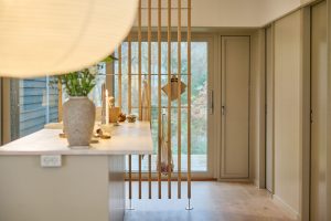 WoodUpp Raumteiler aus schmalen, senkrechten Holzlatten mit runden Metallfüßen trennt Küche und Flur; daran hängen WoodUpp-Querstangen und Haken, an denen Schürzen, Topflappen und eine Netz-Einkaufstasche befestigt sind. Im Vordergrund eine Kücheninsel mit Vase und Kerze, skandinavisch ruhig, in hellem Eicheton und minimalistischem Design.