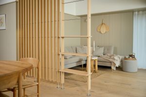 WoodUpp Holzraumteiler mit schlanken, vertikalen Eichenlamellen und integriertem Leiterregal mit drei Holzstufen, Metallquerstreben und verstellbaren Edelstahlfüßen; helles, natürliches Holz im modernen Wohnzimmer, trennt Ess- und Wohnbereich vor grauem Sofa, Teppich und sanfter Dekoration. Neben Sitzsofa, Kissen und beigem Plaid stehen Rattantisch, Kerzenhalter und ein Pouf mit Teekanne.