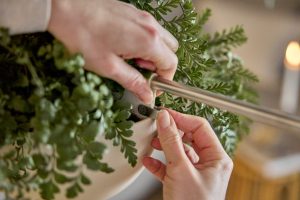 Nahaufnahme: Hände befestigen eine schlanke Edelstahlstange an einem weißen Blumentopf mit grüner Farnpflanze. Zu sehen ist ein kleines schwarzes WoodUpp-Clip am Topfrand, offenbar Teil der WoodUpp-Aufhängung. Man erkennt Erde im Topf und Finger, die ein transparentes Klebeband straffziehen, während im Hintergrund eine Kerze unscharf leuchtet.