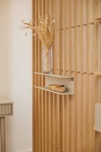 WoodUpp-Vertikale Holzlamellenwand in hellem Holzton mit zwei matten, sandfarbenen WoodUpp-Schweberegalen aus Metall; auf dem oberen Regal eine hohe Glasvase mit getrockneten Gräsern, auf dem unteren zwei kleine handgedrehte Holzschalen. Minimalistisches Design mit feinen senkrechten Lamellen und klaren Linien.