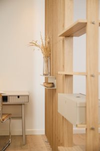 WoodUpp Raumteiler aus hellen Holzlamellen mit integriertem sandfarbenem Metall Wandregal, darauf eine kleine Holzschale und eine Glasvase mit getrockneten Blumen; daneben ein moderner Schreibtisch und Stuhl, sorgfältige Verarbeitung, sichtbare Holzmaserung, skandinavischer Stil, helle Holzböden und dezente Metallakzente, weich ausgeleuchtet, ruhige natürliche Farbpalette.