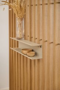 WoodUpp Lamellenwand aus hellem Eichenholz mit vertikalen Lamellen und warmem Ton; daran zwei beigefarbene Metallregalböden von WoodUpp. Auf dem oberen Brett eine hohe Glasvase mit getrockneten Gräsern, auf dem unteren Brett zwei kleine hölzerne Schalen — zurückhaltende, natürliche Dekoration im modernen Stil.