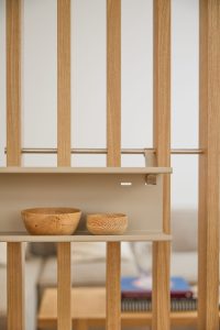 WoodUpp Raumteiler aus senkrechten Eichenlatten mit Edelstahlquerstange und beigefarbener Regalblende: an einer schmalen WoodUpp Ablage stehen zwei gedrechselte Holzschalen, eine größere und eine kleinere, mit sichtbarer Maserung und warmem Ton. Im Hintergrund unscharf Sofa und Bücher, klarer Fokus auf Holz und Ablage.