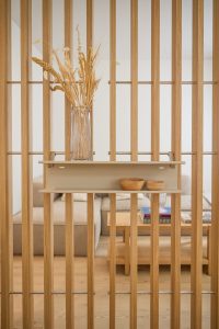 Holztrennwand von WoodUpp mit schlanken, senkrechten Eichenlatten und feinen Metallstreben, an denen eine minimalistische, pulverbeschichtete beige WoodUpp Wandablage als schwebendes Regal und praktische Ablagefläche montiert ist. Darauf zwei kleine Holzschalen und eine hohe Glasvase mit Trockenblumen, vor Sofa und Couchtisch.