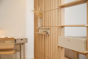 WoodUpp: Helle Szene mit vertikaler Eichen‑Lattenwand, daran ein beiges Wandregal/Schubfach mit zwei kleinen Holzschalen und einer Glasvase mit getrockneten Gräsern. Daneben ein beiger Schreibtisch mit Rattanstuhl sowie offene Regalboxen als dezentes Raumteiler- und Aufbewahrungssystem in warmen Holz- und Sandtönen.