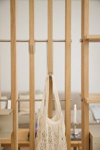 WoodUpp Raumteiler aus heller Eiche mit sichtbarer natürlicher Maserung, vertikalen Lamellen und horizontalen Metallstangen. An den Stangen hängen zwei schlanke WoodUpp-Edelstahlhaken (u‑förmig), einer trägt eine cremefarbene Baumwoll‑Netz‑Einkaufstasche. Rechts ist ein offenes WoodUpp‑Regalbrett sichtbar, im Hintergrund unscharf Kerze und Couch.