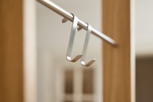 Zwei schlanke WoodUpp-Haken aus gebürstetem Aluminium hängen nah aufgenommen an einer runden Metallstange, U-förmig mit dezent gerundeten Enden und sichtbarem Logo an der Unterseite. Vor verschwommenem Holzrahmen im Hintergrund zeigen die fein satinierte Oberfläche und das minimalistische Design hohe Qualität und Funktionalität.