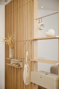 WoodUpp Eichenlamellen-Raumteiler mit sichtbarer Holzmaserung und vertikalen Lamellen, ergänzt durch modulare, integrierte Holzregale und schmale sandfarbene Metallablagen. Metallhaken, eine beige Aufbewahrungsbox und eine Metallstange mit Tassenhaken unterstreichen die minimalistische, modulare Funktion. Dazwischen hängen ein gehäkelter Netz-Einkaufsbeutel, Trockenblumen, Holzschalen und Kerzengläser.