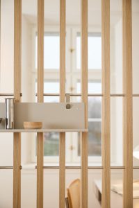 WoodUpp Lamellenwand aus hellem Eichenholz mit vertikalen, schlanken Latten und feiner Maserung, daran montiertes hellgraues Organizer‑Panel/Regal von WoodUpp mit matter Oberfläche, Edelstahlbecher als Halter und kleine gedrehte Holzschale auf der Ablage; dezenter Raumteiler mit Metallstangen, sanftem Tageslicht und klarer, minimalistischer Formsprache.