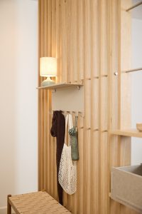 Vertikale WoodUpp-Lamellenwand aus hellem Eichenholz, die Tiefe und warme Atmosphäre schafft, kombiniert mit einem beigefarbenen WoodUpp-Wandboard aus matt lackiertem Metall: flache Ablage über fünf Haken. Auf der Ablage steht eine Lampe, an den Haken hängen Schal, Netzbeutel und zusammengefalteter Regenschirm; davor ein geflochtener Sitzhocker.