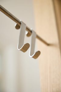 Naheaufnahme zweier schlanker WoodUpp Über-Türhaken aus gebürstetem Metall, nebeneinander an einer goldfarbenen Garderobenstange hängend. Die matte, S‑förmige Form hat abgerundete Enden mit feiner WoodUpp‑Logo‑Gravur; Oberfläche leicht gebürstet, ideal für Mäntel, Handtücher oder Taschen, vor hellem Holzrahmen. Sanftes, warmes Licht akzentuiert die Konturen, minimalistisches Design.
