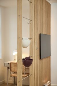 WoodUpp Raumteiler aus hellen Eichenlamellen mit sichtbarer natürlicher Holzmaserung, bestückt mit WoodUpp Hängeelementen: zwei halbrunde Aufbewahrungsschalen (weiß und dunkelviolett) an silbernen Metallstangen und mehreren Haken. Minimalistisches, funktionales Design; im Hintergrund Schreibtisch, geflochtener Stuhl, Lampe und wandmontierter Fernseher, Kerzen auf Ablage.