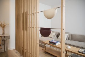 WoodUpp Raumtrenner aus vertikalen Eichenleisten mit eingezogenen Edelstahlstangen, an denen zwei halbrunde WoodUpp Hängekörbe hängen (oben hellbeige, unten dunkelrot). Schlichte skandinavische Wohnzimmereinrichtung mit Sofa, Holz-Couchtisch und Dekovase; die halbkuppelförmigen Körbe bieten Platz für Bücher, Pflanzen oder Kleinteile.