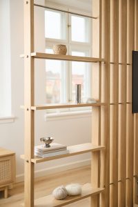 WoodUpp Raumteiler aus hellem, naturbelassenem Eichenholz mit schlanken vertikalen Lamellen, sichtbaren Metallbolzen und feinen Metallstangen sowie drei offenen Regalböden. Auf den WoodUpp-Regalböden: geflochtener Korb, hohe Edelstahlvase, kleiner Dekostein, Bücherstapel mit silberner Schale und zwei Wollknäuel mit Stricknadeln, im hellen skandinavischen Wohnstil, beleuchtet von Tageslicht.