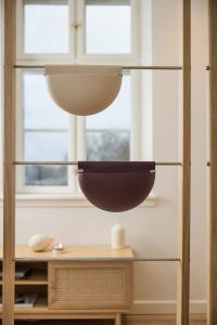 Zwei WoodUpp Hängebehälter hängen in einem schmalen Eichenrahmen an Edelstahlstangen: oben ein sandfarbenes, halbkreisförmiges Wandgefäß, unten ein dunkelrotes/burgunderfarbenes Pendant. Minimalistische, matte Oberfläche und klare Formgebung, beide als platzsparende Pflanz- oder Aufbewahrungsbehälter sichtbar, im skandinavischen Stil arrangiert.