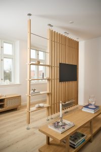 WoodUpp Raumteiler mit vertikalen Eichenlamellen, decken- und bodenmontierten Metallstützen, schlanken Metallfüßen, integrierten offenen Regalböden und TV‑Halterung im Wohnzimmer. Dazu ein schlanker WoodUpp Eichen-Couchtisch und ein Sideboard mit Rattanblende, im natürlichen Eichenfarbton, maßgefertigt und minimalistisch gestaltet.