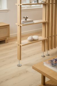 WoodUpp Raumteiler-Regal aus hellem Eichenholz in warmem Ton mit schmalen Regalböden, verstellbaren Metallfüßen und vertikalen Lamellen im minimalistischen Design; darauf Bücher, eine kleine silberne Schale, Wollknäuel und eine Stricknadel. Im Hintergrund ein WoodUpp Lowboard mit Wiener Geflecht sowie Teil eines WoodUpp Couchtisches.