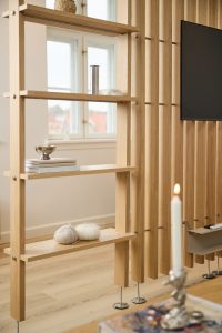 WoodUpp Raumteiler aus hellem, naturbelassenem Eichenholz mit senkrechten Lamellen und integrierten, freitragenden WoodUpp-Regalböden. Das schlanke Trennmöbel steht auf Metallfüßen, trägt Bücher, ein Schälchen und Wollknäuel; rechts ist ein wandähnliches Lamellenfeld mit TV zu sehen, das Licht und Offenheit bringt.