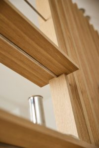 WoodUpp Lamellen‑Raumteiler und Regalboden aus naturbelassener Eiche, Großaufnahme senkrechter Holzlatten mit horizontalem Brett, verbunden durch schlanke Edelstahlstangen; feine Holzmaserung, präzise gefaste Kanten, warmes Holzfinish, sichtbarer zylindrischer Metallfuß, modernes, filigranes Design und sorgfältige Verarbeitung, ideal als Raumteiler, Regal oder dekoratives Element im Wohn‑ oder Bürobereich.