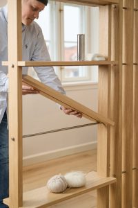 WoodUpp Regal-Raumteiler aus hellem, naturbelassenem Eichenholz mit senkrechten Lamellen, verstellbaren Ablageböden und integrierten Edelstahlstangen; ein Mann stellt einen schräg einstellbaren Einlegeboden ein. Auf den Böden liegen zwei Knäuel Wolle mit Stricknadel, eine Seife und ein Edelstahlbecher im typischen minimalistischen, modularen Design.