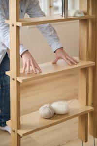 WoodUpp-Leiterregal aus massiver Eiche mit geölter Oberfläche, sichtbarer Holzmaserung, klaren flachen Regalbrettern und sichtbaren Metallbolzen; eine Person in gestreiftem Hemd richtet mit beiden Händen ein mittleres Fach. Auf dem unteren Brett liegen zwei Wollknäuel und Stricknadeln, oben ein glatter Dekostein.