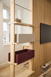 WoodUpp Raumteiler aus hellem Eichenholz mit Regalleisten und sichtbaren Metallbolzen, bestückt mit zwei WoodUpp Filz-Boxen in Grau und Bordeaux sowie einer kleinen Ablage mit Garnrollen. Mit natürlicher Oberfläche. Im Hintergrund Holzlamellenwand mit TV, Fensterlicht und Bürobereich, moderne, luftige Raumaufteilung.