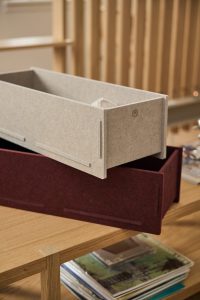 Auf dem Bild zwei WoodUpp-Boxen aus robustem Filz: eine hellbeige Box mit dezenter WoodUpp-Logo-Prägung liegt über einer weinroten Filzschublade. Beide rechteckigen, modularen Aufbewahrungsbehälter stehen auf einem Eichenholz-Tisch; darunter ist ein Stapel Magazine zu erkennen, mit feinen Einfassungen und Steckverbindungen, skandinavischem, minimalistischem Design.