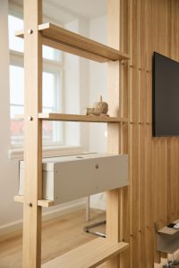 WoodUpp Raumteiler Regal aus hellem Eichenholz mit schlanken vertikalen Stützen und offenen Regalböden, darauf ein eleganter grauer rechteckiger WoodUpp Kabelkasten zur Kabelführung sowie dekorative Garnrollen. Rechts ist eine Holzlamellenwand mit Fernseher zu sehen — moderne, minimalistische modulare Wohnraumlösung von WoodUpp.