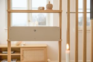 WoodUpp Raumteiler aus hellem Eichenholz mit schlanken Lamellen und offener Regalstruktur, oben zwei Garnrollen; mittig das beigefarbene WoodUpp Technikmodul/Verblendung mit dekorativer Lüftungsöffnung; im Hintergrund ein Sideboard mit geflochtener Tür, Kerzen und warmem, stimmungsvollem Licht, vorne eine brennende Kerze im Fokus, neutrale, skandinavisch-minimalistische Einrichtung und weiches Tageslicht durch Fenster.