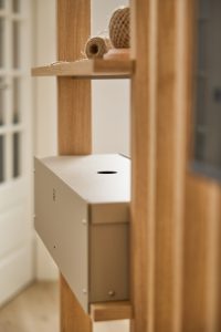 Nahe Aufnahme eines WoodUpp‑Regals aus Eichenholz mit schlanken, vertikalen Streben und hellen Lamellen. Eingebaut ist eine matte, beige‑metallic Einschubbox/Schublade mit rundem Öffnungsloch, sichtbaren Schrauben und kleinem Löchermuster. Auf dem oberen Regalbrett liegen zwei Rollen Jute‑Garn, natürliches Licht betont Materialstruktur und Ästhetik.
