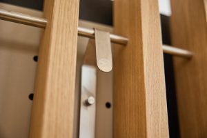 Detailaufnahme eines WoodUpp-Garderobenmoduls: zwei vertikale Eichenholzlamellen in natürlichem hellbraun mit sichtbarer Maserung, eine matt gebürstete Edelstahlstange und ein schlanker Metallclip/Haken mit abgerundetem Ende und dezent eingeprägtem WoodUpp-Logo. Im Hintergrund eine Metallaufhängung mit Rundstift; die Nahperspektive betont die warme Optik und feine Verarbeitung.