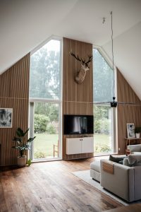 Modernes Wohnzimmer mit hohen Fenstern und WoodUpp-Lamellenwandpaneelen aus hellem Holz. Sichtbar sind WoodUpp-vertikale Wandverkleidungen hinter TV und Fenster, ein WoodUpp-TV-Sideboard mit Holzfront und eine kleine WoodUpp-Armlehnablage aus Lamellen auf dem Sofa, ergänzt durch Parkettboden, Dekor und Pflanzen.