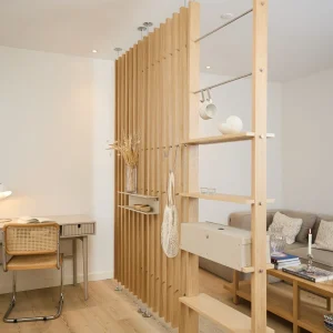 WoodUpp Raumteiler aus hellen, vertikalen Eichenlamellen trennt Wohn- und Arbeitsbereich; daran befestigt sind WoodUpp offene Regalbretter, eine Edelstahlstange mit Tassenhaken, mehrere Metallhaken, eine beige klappbare Aufbewahrungsbox sowie eine aufgehängte Netz-Einkaufstasche und Vase mit Trockenblumen in natürlichem Holzton und stabilem Profil.