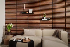 Wohnzimmer mit eleganter WoodUpp-Holzlamellenwand in warmem Nussbaumton und feinen schwarzen Fugen sowie zwei schwarzen WoodUpp-Wandregalen, bestückt mit Kerzenständer, Buch und kleiner Vase. Vor der Wand ein beiges Ecksofa mit Kissen, Tulpen in Glasvase und Tablett mit Tassen vervollständigen die ruhige, moderne Einrichtung.