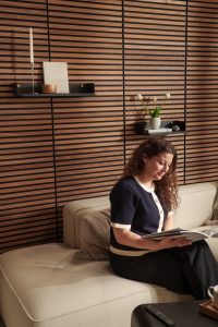 WoodUpp-Wandverkleidung aus horizontalen dunklen Holzlamellen bildet den Hintergrund; zwei WoodUpp schmale, schwarz lackierte Wandregale mit sichtbaren Messingschrauben und minimalistischer Optik tragen Kerzen, Bücher, eine Holzschale und eine Vase mit Tulpen; im Vordergrund sitzt eine Frau auf einem hellbeigen Sofa und liest.