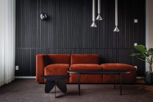 Modernes Wohnzimmer mit WoodUpp vertikalen Lamellenpaneelen in mattschwarz, die die gesamte Wand mit schmalen, gleichmäßigem Abstand strukturieren. Davor ein rostrotes, modulares Samtsofa, zwei schwarze Beistelltische (oval und dreiblättrig), hängende Metalllampen und eine Zimmerpflanze. WoodUpp betont Tiefe, Optik und Akustik im minimalistischen Stil.