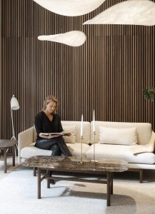 Moderne Sitzecke vor einer WoodUpp Lamellenwand aus dunklem Holz: feine, vertikale Lamellen bilden eine gleichmäßige Textur. Helles Stoffsofa mit Kissen, ovaler Marmor-Couchtisch auf Holzbeinen, zwei schlanke Messing‑Kerzenhalter, weiße Stehlampe und eine Frau, die entspannt eine Zeitschrift liest und schaffen ein warmes, einladendes Ambiente mit natürlicher Farbpalette.