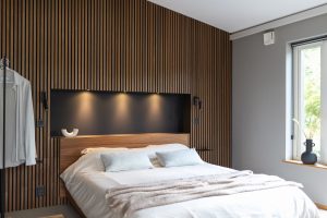 Modernes Schlafzimmer mit markanter WoodUpp-Holzlamellenwand in warmem Nussbaumton und dazugehörigem WoodUpp-Holzkopfteil mit eingelassener, dunkel hinterlegter Nische und drei Spotleuchten. Weiß bezogenes Bett mit hellgrauen Kissen, beige Decke, schwarze Wandleselampen, Fenstersims mit dunkler Vase und Tageslicht von rechts, minimalistisches Design.