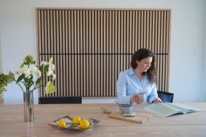 Eine Frau sitzt am hellen Esstisch vor einer großen WoodUpp-Wandverkleidung aus vertikalen Eichenlamellen mit schwarzem Hintergrund und schlankem Rahmen; auf der Tischplatte liegen ein Holz-Schneidebrett, Keramikbecher, offene Zeitschrift, eine Schale mit Zitronen und eine Vase mit weißen Blumen im reduzierten, skandinavisch inspirierten Stil.