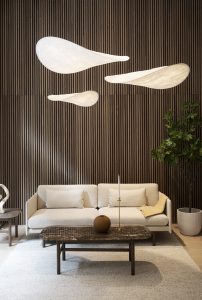 Modernes Wohnzimmer mit WoodUpp-Holzlamellen-Wandpaneelen in dunklem Nussbaumton als markantem Hintergrund. Drei schwebende, organisch geformte weiße Hängeleuchten, ein beiges Sofa mit Kissen, niedriger dunkler Holztisch mit runder brauner Vase, schlanker Kerzenhalter und große Topfpflanze; die WoodUpp-Lamellen betonen vertikale Struktur.