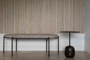 Schlichte Szene mit WoodUpp-Lamellenwand aus hellen, vertikal angeordneten Holzlatten und grauem Filzzwischenraum, gleichmäßig strukturiert. Dazu eine WoodUpp-Eingangsbank mit dünnem schwarzen Metallgestell und gepolsterter, beige-melierter Sitzfläche. Rechts ein kleiner schwarzer Beistelltisch mit runder Platte und marmoriertem, bauchigem Fuß auf hellem Parkettboden vor weißem Sockel in minimalistischem Stil.