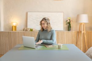 Frau sitzt am hellen, minimalistischen Tisch und arbeitet am Laptop auf einer grünen WoodUpp Schreibtischunterlage; daneben Notizbuch und Tasse. Im Hintergrund ein WoodUpp Sideboard mit vertikalen Holzlamellen und eine helle WoodUpp Stehlampe mit dreibeinigem Holzgestell, dekoriert mit Vase und Bild.