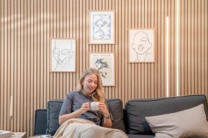 Junge Frau sitzt auf dunkelgrauem Sofa vor einer WoodUpp-Holzlamellenwand. Sichtbare WoodUpp-Vertikallamellen in hellem Eichenholz, gleichmäßig angeordnete, feine Maserung. Vier schmale WoodUpp-Bilderrahmen mit hellen Holzleisten und minimalistischen Kunstdrucken. Warme LED-Streifen rechts, Kerzenleuchter links, kuschelige Decke und Kaffeetasse in den Händen und natürliche Farbstimmung.